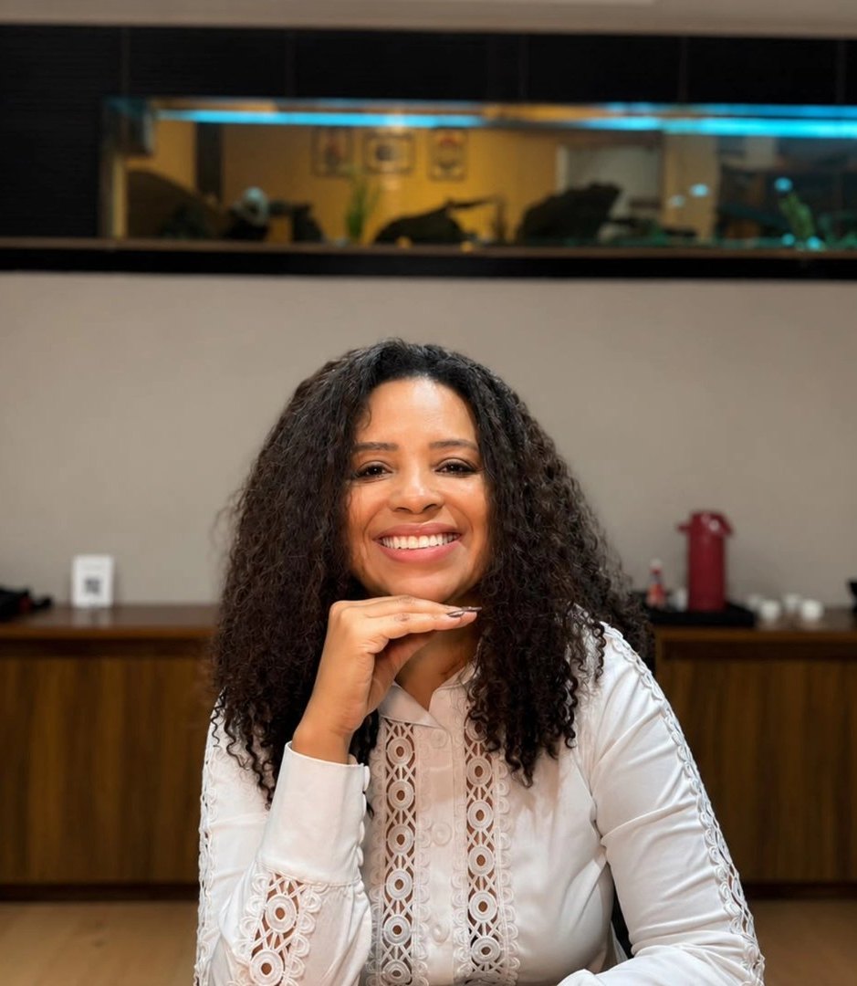 Viviane Tobias sorrindo, com a mão apoiada no queixo, de blusa branca